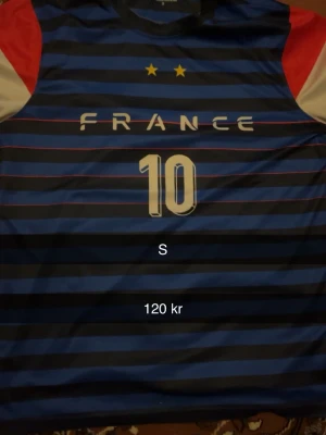 Frankrike fotbollströja blå S - Säljer en blå och svart randig fotbollströja med FRANCE-tryck och nummer 10 på bröstet. Tröjan har röda och vita detaljer på axlarna samt två gula stjärnor ovanför texten. Perfekt för dig som gillar fotboll och vill stötta Frankrike.