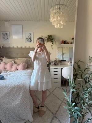 Vit volangklänning med broderi - Söt vit kortklänning med volangärmar och smock i midjan. Klänningen har broderade detaljer och en luftig, lätt känsla. Perfekt för varma dagar och har en feminin siluett med dubbla volanglager nertill.