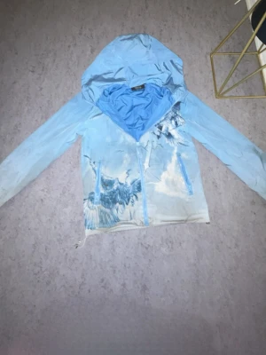 Blå vindjacka från Trapstar med tryck size s - Snygg ljusblå vindjacka från Trapstar med huva och dragkedja. Jackan har ett unikt tryck med vingar och moln på framsidan. Tillverkad i lätt och glansigt material, perfekt för streetwear-stil. Jackan har dragsko nertill för justerbar passform.