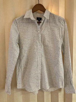 Vit prickig skjorta från Gant - Snygg vit skjorta från Gant med små svarta prickar över hela tyget. Skjortan har klassisk krage, knäppning framtill och långa ärmar. Tillverkad i ett tunt och skönt material som passar perfekt för lager på lager under vintern.