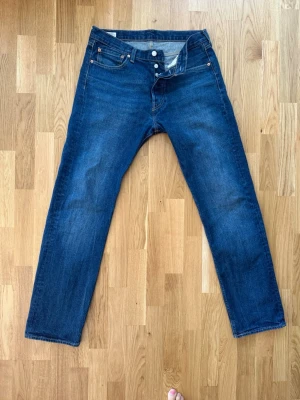 Blå raka jeans från Levi's 501 - Klassiska blå jeans från Levi's med rak passform och fem fickor. Jeansen har knappgylf och tydliga sömmar på bakfickorna. Perfekta för en avslappnad och tidlös stil. Materialet är robust denim i bomull. 33/34
