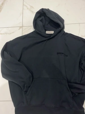 Svart Essentials Fear of God hoodie - Säljer en svart hoodie från Essentials Fear of God med tryck på bröstet och stor logga på ryggen. Hoodien har en klassisk huva och magficka framtill. Tillverkad i mjukt bomullsmaterial som är perfekt för en avslappnad streetwear-look.