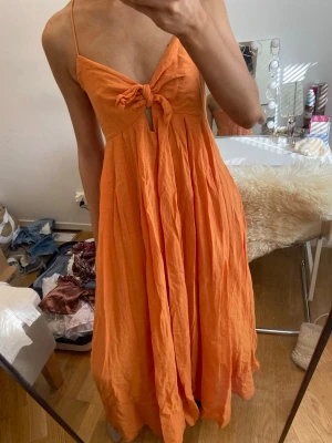 Orange långklänning med knyt framtill - Snygg orange långklänning med smala axelband och knyt framtill. Klänningen har en luftig passform och är tillverkad i ett lätt material som passar perfekt för varma dagar. Den har en liten öppning under bysten och faller fint längs kroppen.