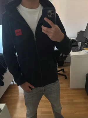 Svart hoodie med dragkedja från HUGO - Snygg svart hoodie från HUGO med dragkedja framtill och röd logotyp-patch på bröstet. Hoodien har justerbar huva med snören och två fickor framtill. Tillverkad i mjukt material som känns skönt mot huden. Perfekt för en avslappnad och stilren look.