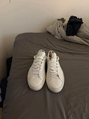 Vita common projects  - Säljer ett par stilrena vita common projects,  med en låg profil och vita snören. Skorna har en enkel och clean design som passar till det mesta. Perfekta för dig som gillar en tidlös och fräsch look erbjud annars pris om du vill ha ett lägre .