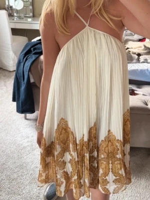 Pleated halter summer dress - Superfin plisserad kortklänning från H&M i vitt med beige paisleymönster nertill. Klänningen har tunna axelband och luftig passform, perfekt för varma dagar. Materialet känns lätt och flowy, och mönstret ger en bohemisk vibe.