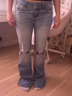 Ljusblå bootcut jeans med slitningar - Säljer ett par ljusblå bootcut jeans med slitna knän och utsvängda ben. Jeansen har klassiska fem fickor, dubbla knappar i midjan och dekorativa bakfickor med lock. Perfekta för en avslappnad och trendig look.
