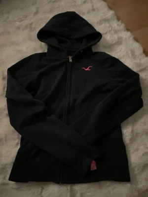 Svart hoodie från Hollister XS - Svart zip-hoodie från Hollister med huva och dragkedja framtill. Klassisk design med rosa Hollister-logga på bröstet och rosa detaljer på insidan av kragen. Perfekt för en avslappnad och stilren look. Lappen vid ärmen har lossnat lite grann (se på bild 3) Pris kan diskuteras 