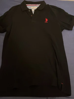 Svart pikétröja från U.S. Polo Assn - Svart pikétröja från U.S. Polo Assn med röd logga broderad på bröstet. Klassisk krage och knappslå framtill. Slim fit-modell som passar perfekt till jeans eller shorts. Skön bomullskvalitet och stilren design.
