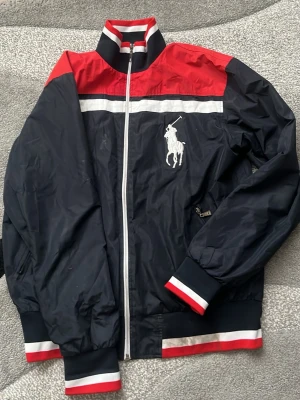 Ralph Lauren vindjacka i navy och rött - Snygg vindjacka från Ralph Lauren i marinblått med röda och vita detaljer. Jackan har dragkedja, ribbade muddar och stor vit logga på bröstet. Materialet är lätt och vindtätt, perfekt för blåsiga dagar. Klassisk sportig look med meshfoder på insidan.