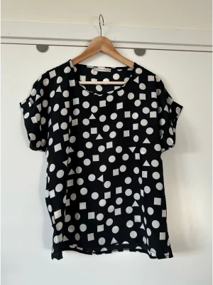 Svart och vit kortärmad blus / topp - storlek M - Superfin svart och vit kortärmad topp blus med ett geometriskt mönster av cirklar, kvadrater och trianglar. Liva Girl, storlek M. Toppen har korta ärmar med en liten uppvikt kant och en rundad halsringning. Passar bra för en avslappnad men stilfull look. Tunt och följsamt material. Mycket gott skick, som ny!