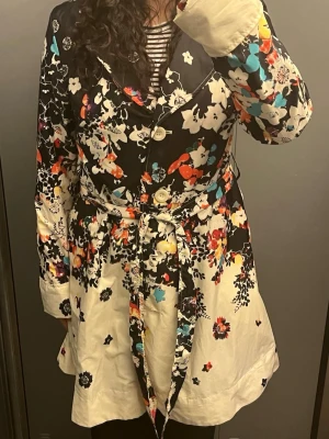 Blommig trenchcoat med knytband - Snygg trenchcoat med färgglatt blommönster i svart, vitt, rött, blått och orange. Jackan har stora vita knappar, klassisk krage och ett matchande knytband i midjan. Materialet ser ut att vara bomull eller siden och modellen är A-linjeformad.