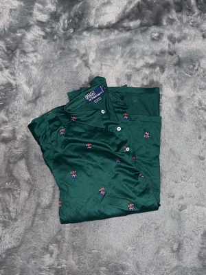 Polo Ralph Lauren All-Over Crest Embroidery - En extremt snygg mörkgrön pikétröja från Polo Ralph Lauren med ett sällsynt och detaljrikt mönster. Tröjan är helt täckt med små broderade vapensköldar (crests). Toppskick 