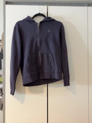 Mörkblå hoodie från GANT - Mörkblå hoodie med dragkedja från GANT. Klassisk modell med huva och två fickor framtill. Liten broderad GANT-logga med sköld på bröstet. Skön och stilren, perfekt till jeans eller joggers.