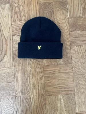 Svart mössa från Lyle & Scott - En svart ribbstickad mössa från Lyle & Scott med uppvikt kant och den klassiska gula örnloggan broderad framtill. Oanvänd.