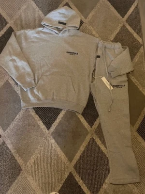 Grått loungewear-set från Essentials - Mysigt grått loungewear-set från Essentials med hoodie och matchande byxor. Hoodien har magficka och tryckt logga på bröstet, byxorna har dragsko i midjan och logga på benet. Perfekt för chill dagar hemma.