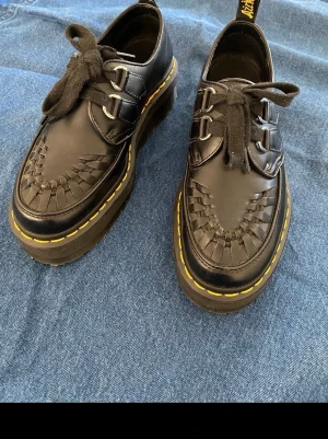 Svarta Dr. Martens loafers med platåsula - Säljer ett par svarta loafers från Dr. Martens med grov platåsula och klassisk gul söm. Skorna har flätad detalj framtill och är gjorda i läder. Perfekta för dig som vill ha en edgy och stilren look.