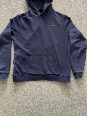 Mörkblå hoodie från Lyle & Scott - Snygg mörkblå hoodie från Lyle & Scott med klassisk gul logga på bröstet. Hoodien har huva med dragsko och en stor magficka framtill. Tillverkad i mjuk bomullsmix som är skön att ha på sig. Perfekt för en chill och stilren look.