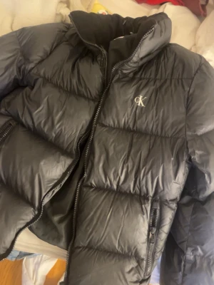 Svart pufferjacka från Calvin Klein - Svart pufferjacka från Calvin Klein Jeans med hög krage och dragkedja framtill. Jackan har två sidofickor med dragkedja och diskret CK-logga på bröstet. Perfekt för kalla vinterdagar och har en klassisk, stilren look. (utan luvan) 