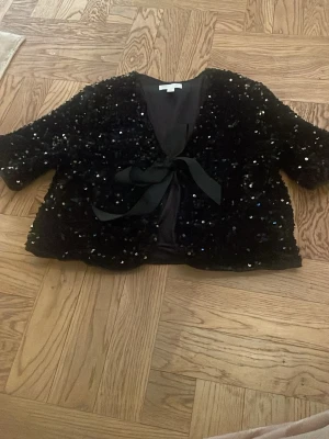 Svart paljettkofta med rosett - Glittrande svart kofta täckt av paljetter med korta ärmar och en rosett framtill. Koftan har en öppen design utan knappar. Perfekt för dig som vill sticka ut och addera extra bling till din outfit på en dag när man vill klä sig lite extra fint.