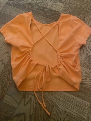 Orange croppad topp med öppen design på ryggen - Trendig orange croppad topp med korta ärmar och snygg öppen rygg med snörningsdetalj. Toppen har en rundad halsringning och är tillverkad i ett mjukt, stretchigt material som känns skönt mot huden. Perfekt för dig som vill sticka ut med en färgglad och unik look. Färgen är lite starkare än vad den ser ut att vara på bilden tycker jag.