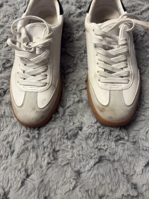 Vita sneakers med beige detaljer - Snygga vita sneakers med beige mockadetaljer och gummisula. Skorna har klassisk låg profil, vita skosnören och stilren design. Perfekta för en clean och trendig look. Materialmix av läder och mocka ger en schysst kontrast. Några skavanker. Se bilder för mer info. Tror att storleken är 37. Jag har storlek 38 och de är för små för mig. 