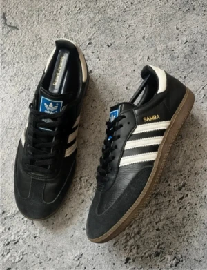 Adidas Samba svarta sneakers - Klassiska Adidas Samba sneakers i svart läder med vita detaljer och de ikoniska tre ränderna på sidorna. Skorna har gummisula och blå logga på plösen. Perfekta för en sportig och stilren look.