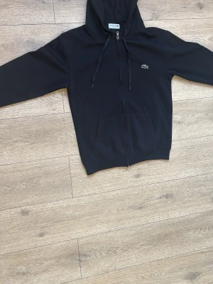 LACOSTE ZIP-HOODIE - SVART ZIP-HOODIE FRÅN LACOSTE MED SVARTA SNÖRREN SILVER DRAGKEDJA GRÖN RÖDA SMÅ DETALJER STORLEK M PASSAR ÄVEN OM MAN HAR S