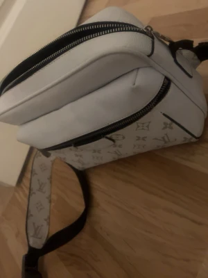 Vit axelväska från Louis Vuitton - Snygg vit axelväska från Louis Vuitton med klassiskt monogrammönster i grått. Väskan har två dragkedjefack och justerbar axelrem med logga. Tillverkad i canvas med svarta detaljer och metallspänne. Perfekt för dig som vill ha en trendig och praktisk väska.
