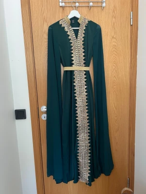 Mörkgrön långklänning med guldiga detaljer - Elegant mörkgrön långklänning från shein med långa ärmar och vackra guldiga broderade detaljer längs hela framsidan och kragen. Klänningen har ett matchande guldigt bälte i midjan och en rak, flytande passform.