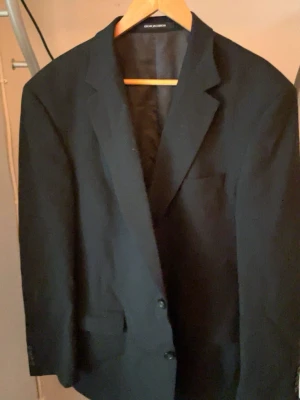 Svart kavaj från Oscar Jacobson - Klassisk svart kavaj från Oscar Jacobson med två knappar framtill och diskreta fickor. Snygg passform och helfodrad insida. Perfekt för en stilren look. Materialet ser ut att vara ullblandning.