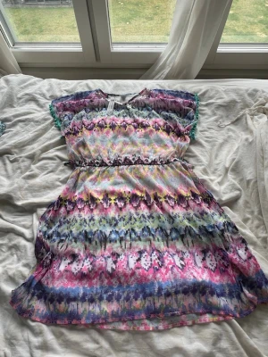 Färgglad kort klänning från Lindex - Superfin kort klänning från Lindex med färgstarkt mönster i rosa, blått, lila och grönt. Klänningen har paljettdetaljer vid halsen, korta ärmar och ett tunt band i midjan. Perfekt för dig som gillar färg och glitter! Aldrig använd💕