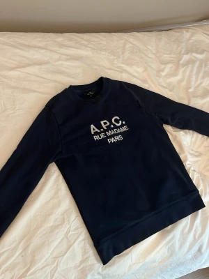 Mörkblå sweatshirt från A.P.C. - Mörkblå sweatshirt från A.P.C. med vit text 'A.P.C. RUE MADAME PARIS' på bröstet. Tröjan har rund halsringning, långa ärmar och är gjord i ett mjukt material som känns skönt mot huden. Enkel och stilren design som passar till det mesta.