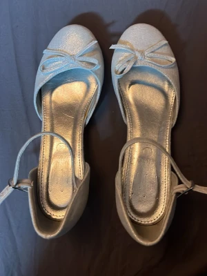 Silvriga ballerinaskor med rosett - Snygga silvriga ballerinaskor med glittrig finish och söt rosett framtill. Skorna har en smal ankelrem med spänne och rundad tå. Perfekta för dig som vill ha en stilren och trendig look. Materialet ser ut att vara syntet. Använda några få gånger 
