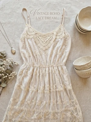 Vintage boho spetsklänning - Romantisk vintageinspirerad boho-klänning i ljus cremevit nyans med tunna axelband och vackra spetsdetaljer. Klänningen har broderade blommor och volanger, samt en omlottliknande spets vid urringningen. Perfekt för sommarens alla tillfällen.