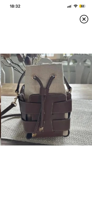 Brun och beige bucket bag med snörning - Snygg bucket bag i brunt skinn med beige tyginsats och snörning upptill. Väskan har justerbar axelrem och detaljer i guld. Perfekt storlek för det viktigaste och en modern design med kontrastsömmar.