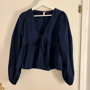 Mörkblå blus med v-ringning - Mörkblå blus från Nelly med v-ringning och dekorativa knappar framtill. Blusen har lång ärm med lätt puff och rynkade detaljer under bysten för en avslappnad och trendig look. Tillverkad i ett mjukt bomullstyg. Som bilderna visar så har några av ”banden” som knapparna fästs i lossnat.