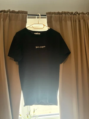 Svart Palm Angels t-shirt - Svart t-shirt från Palm Angels med vit logotyp på bröstet och stor vit text på ryggen. Klassisk rund hals och korta ärmar. Materialet är mjukt och känns som bomull, perfekt för dig som gillar streetwear och enkel design.