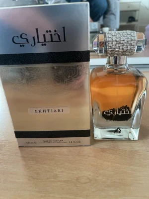 Lattafa Ekhtiari Eau de Parfum 100ml - Ekhtiari Eau de Parfum från Lattafa i en elegant, genomskinlig glasflaska med silverfärgad, mönstrad kork. Doften kommer i en lyxig silverfärgad förpackning med svarta detaljer. Flaskan rymmer 100 ml och har en exklusiv och modern design. Endast testad