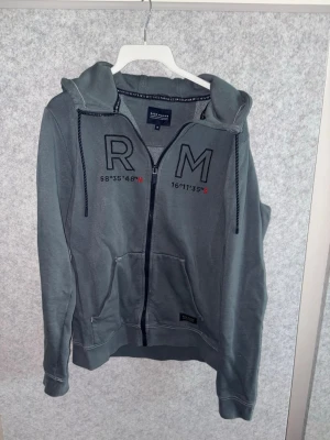 Race marin hoodie mörkblå - Snygg grå hoodie från race marine med dragkedja, huva och fickor framtill. Stora bokstäver 'RM' och koordinater tryckta på bröstet. Tillverkad i mjukt bomullsmaterial och har ribbade muddar vid ärmar och nederkant. Perfekt för en chill och avslappnad stil.