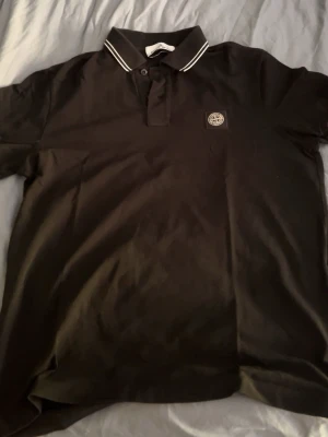 Svart polotopp från Stone Island - Äkta pikétröja från Stone Island med klassisk krage och knappslå. Tröjan har vit detalj på kragen och ikonisk logotyp på bröstet. Perfekt för en stilren och sportig look. 
