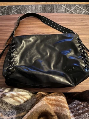 Svart axelväska från Desigual  - Snygg svart axelväska från Luzgral i glansigt konstläder. Väskan har coola snörningsdetaljer längs sidorna och en justerbar axelrem med nitar. Baksidan har en praktisk dragkedjeficka. Perfekt för dig som gillar edgy och trendig stil.
