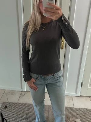 Mörkgrå tröja med knappdetaljer - Snygg skön mörkgrå tröja med rund halsringning och långa ärmar. Tröjan har guldfärgade knappar vid ärmsluten. 