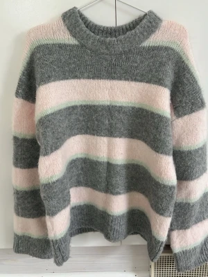 Randig stickad tröja Gina Tricot - Mysig stickad tröja från Gina Tricot i storlek XS. Tröjan har breda ränder i grått, ljusrosa och en tunn mintgrön linje. Den har rund halsringning, lång ärm och är gjord i ett fluffigt, mjukt material som känns riktigt skönt.