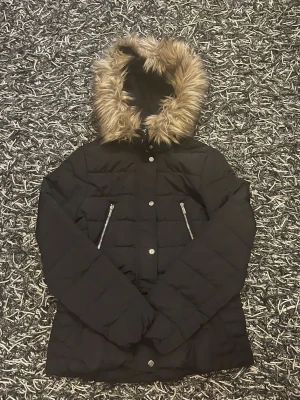 Svart pufferjacka med fuskpäls - Säljer en svart pufferjacka med huva och fuskpälsdetalj. Jackan har två snedställda fickor med dragkedja och stängs med tryckknappar framtill. Perfekt för kalla vinterdagar och har en klassisk, stilren look.