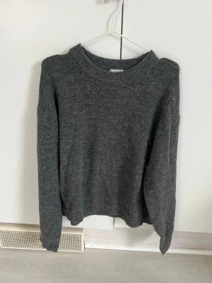 Mörkgrå stickad tröja från H&M - Mysig mörkgrå stickad tröja från H&M med rund halsringning och långa ärmar. Tröjan har en enkel, klassisk design och är perfekt för lager på lager. Materialet känns mjukt och bekvämt mot huden.