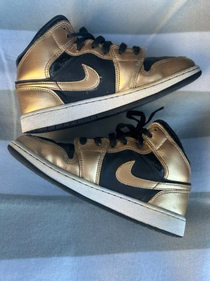 Air Jordan 1 Mid svart/guld - Snygga Air Jordan 1 Mid sneakers i svart och metallic guld. Skorna har klassisk Nike Swoosh på sidan, svarta skosnören och vit mellansula. Tillverkade i läder med ikonisk Air Jordan-logga vid ankeln. Perfekta för dig som vill sticka ut med stilrena detaljer.