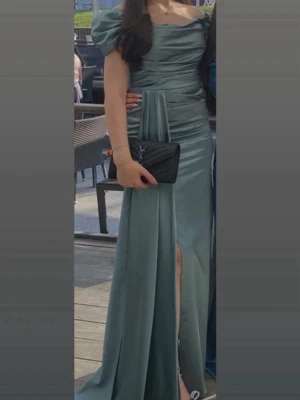 Lång blå satinklänning med puffärm - Elegant långklänning i glansigt blått satinmaterial med puffiga kortärmar och draperad överdel. Klänningen har en markerad midja, slits framtill och v-ringad urringning. Perfekt för bal eller festliga tillfällen.