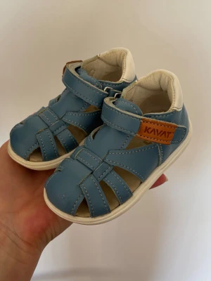 Blåa sandaler från Kavat - Säljer ett par blåa sandaler från Kavat i mjukt läder med vita detaljer och kardborreknäppning. Sandalerna har öppen tå och häl, samt en brun logotyp på sidan. Perfekta för varma dagar och riktigt sköna att ha på sig.