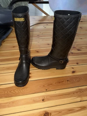 Svarta quiltade stövlar Barbour - Snygga svarta höga stövlar från Barbour med quiltat mönster och spänne vid ankeln. Tillverkade i gummi som tål regn och smuts, perfekta för blöta dagar. Klassisk logga upptill och robust sula för bra grepp.
